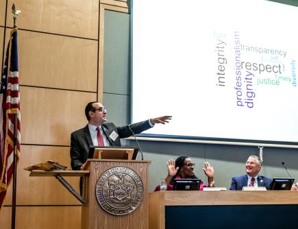 Photo of one judge standing at a podium with two judges seated nearby. Behind them is a large screen with the words Respect and Dignity on it.
